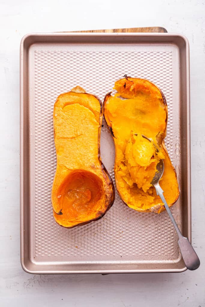 Overhead view of two halves of roasted butternut squash on sheet pan with spoon scooping flesh