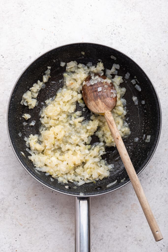 Overhead view of softened onions in skillet with wooden spoon