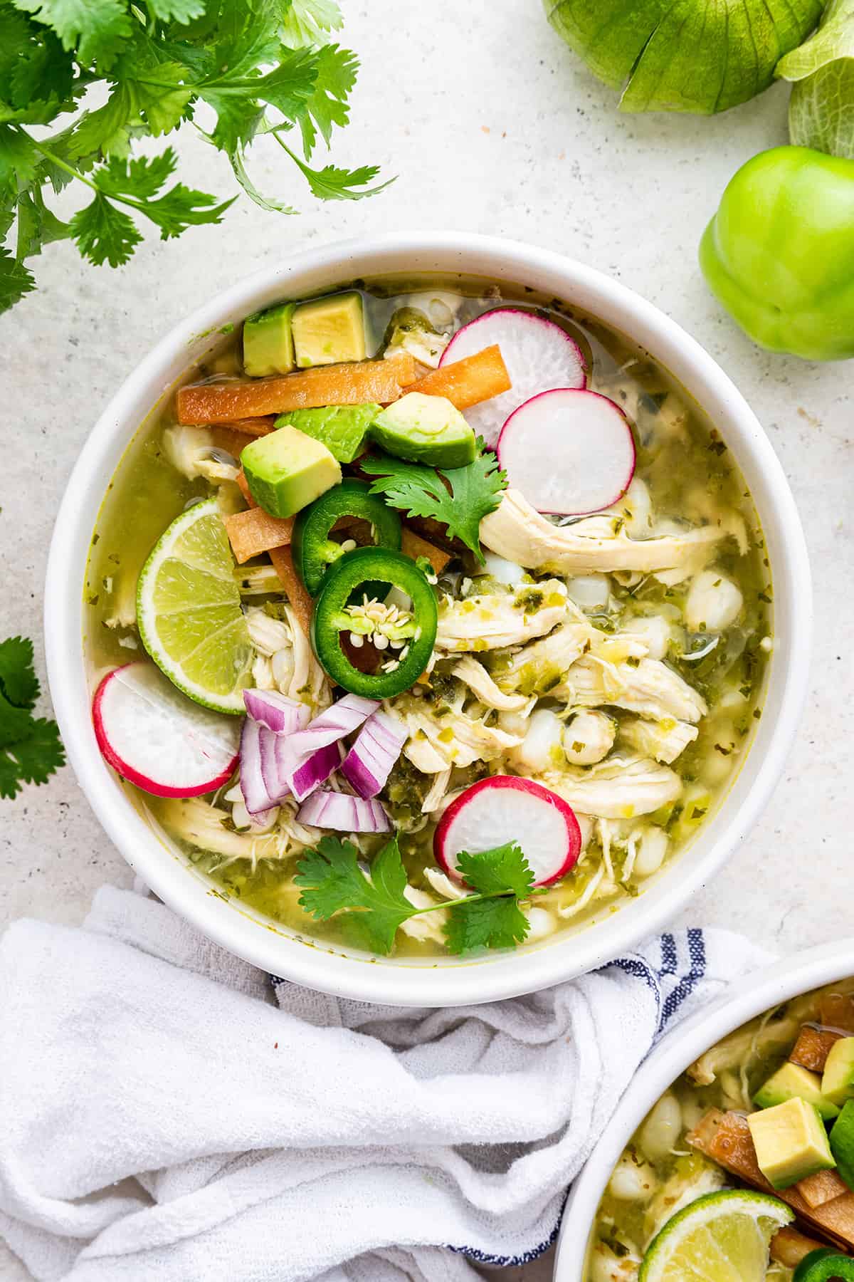 Overhead view of pozole verde in bowl, topped with sliced jalapeños, radishes, lime wedges, avocado, cilantro, and tortilla strips