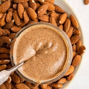 Overhead view of a jar of pumpkin spice almond butter with a spoon submerged in it, sitting on a plate covered in almonds