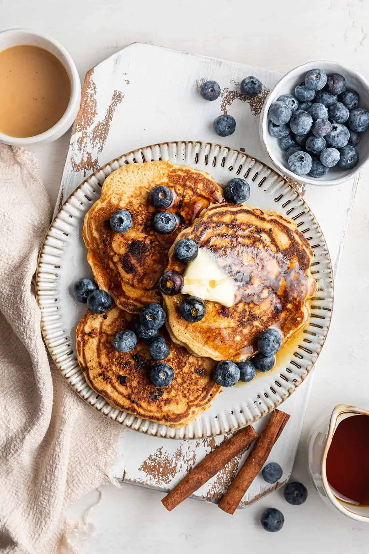 Overhead view of a plate of three pancakes topped with butter, maple syrup, and blueberries, next to a bowl of blueberries, cinnamon sticks, and a cup of coffee