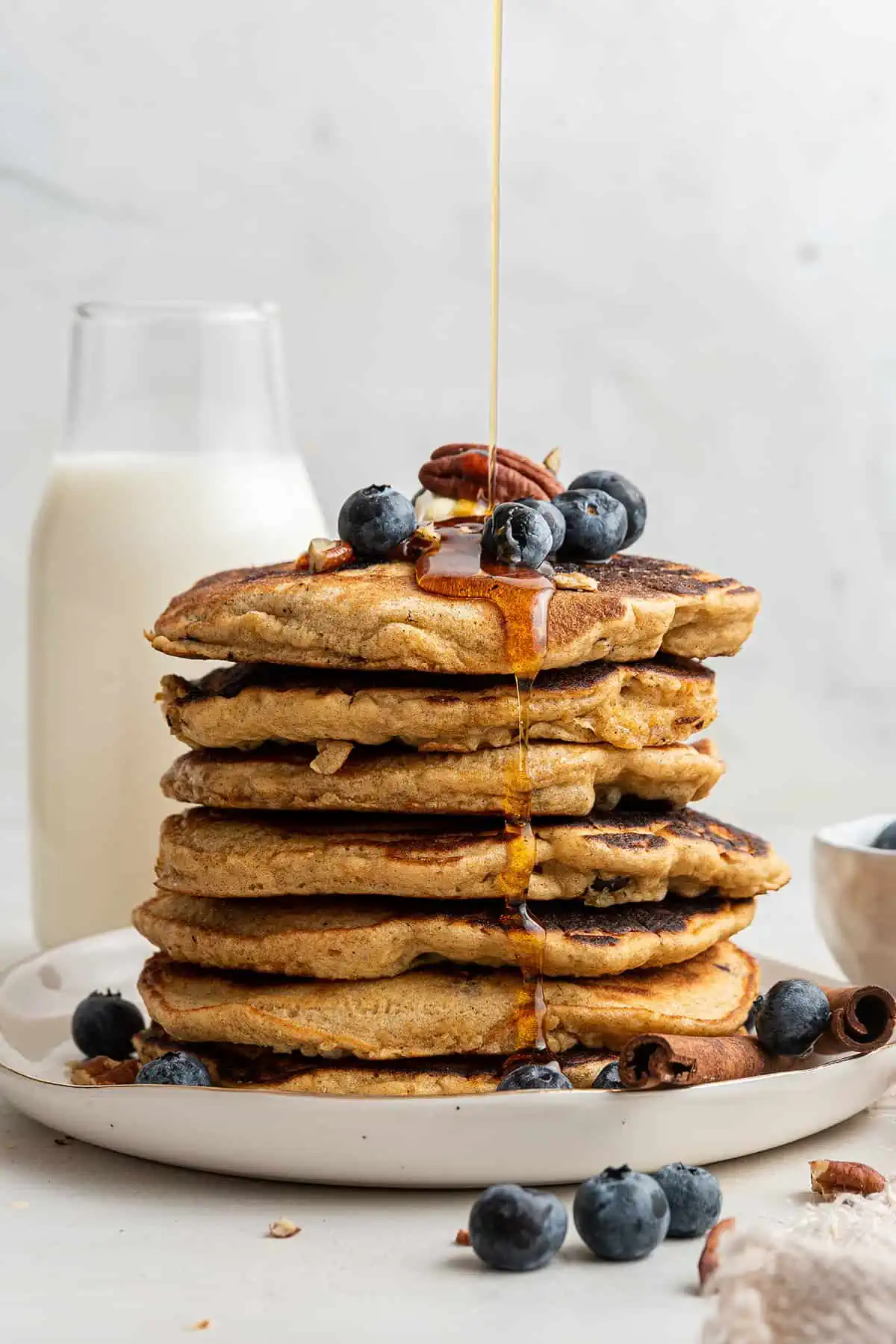 A stack of seven blueberry pancakes, topped with butter, blueberries, and a pecan, with maple syrup being drizzled over it, and a bottle of milk in the background