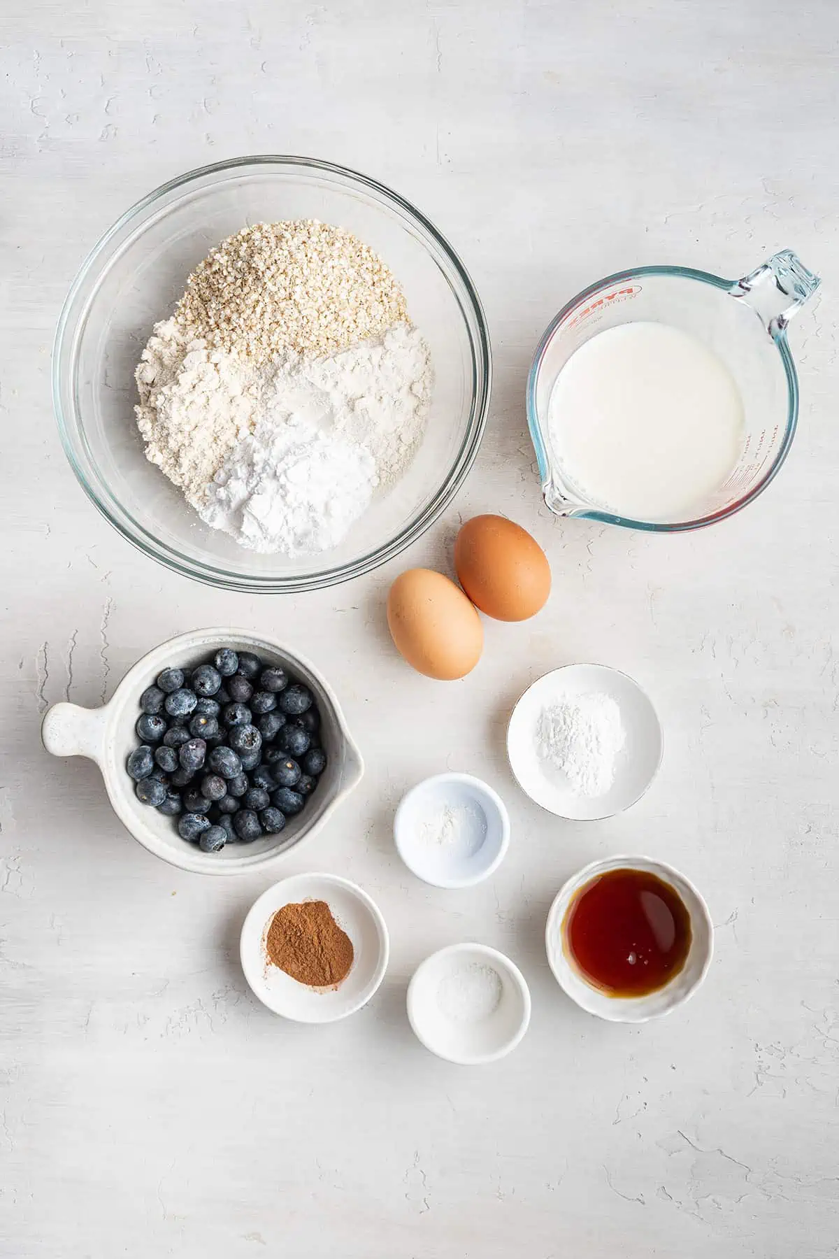 Overhead view of the ingredients needed for gluten-free blueberry pancakes: a bowl of sorghum flour, brown rice flour, tapioca flour, and quinoa flakes, a pyrex of almond milk, a bowl of blueberries, a bowl of maple syrup, a bowl of cinnamon, a bowl of xanthan gum, a bowl of salt, a bowl of baking powder, and two eggs