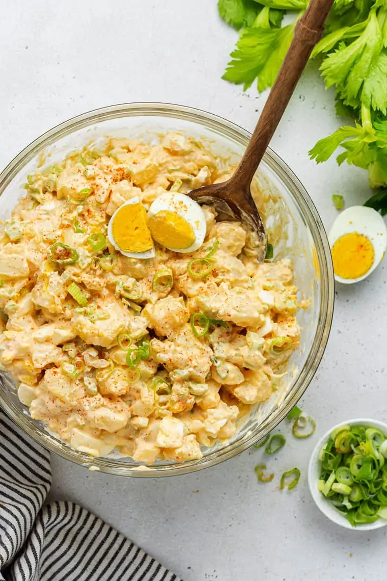 Overhead view of a bowl of potato salad topped with scallions, paprika, and a boiled egg, with a wooden spoon in it, next to a bowl of scallions, half a boiled egg, and some celery