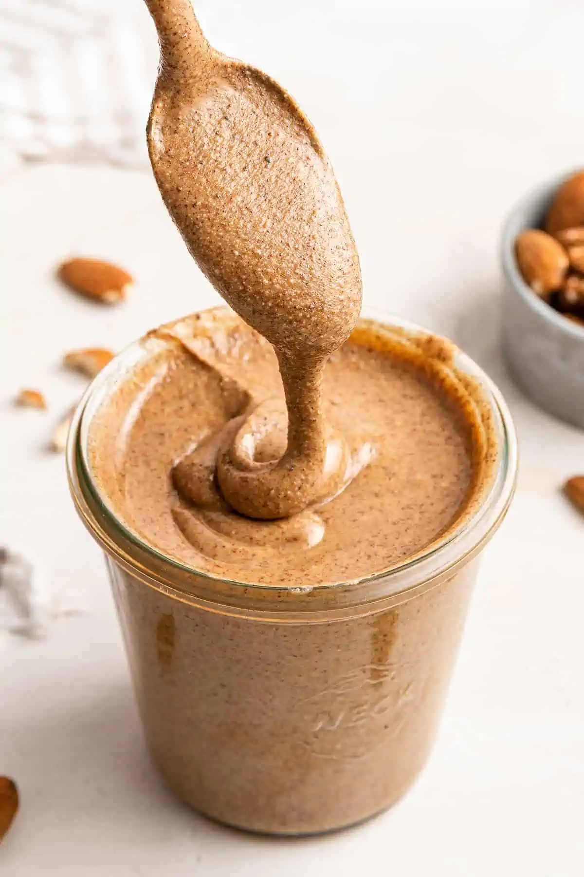 A spoonful of pumpkin spice almond butter drizzling the almond butter into a full jar of it, with a bowl of almonds in the background