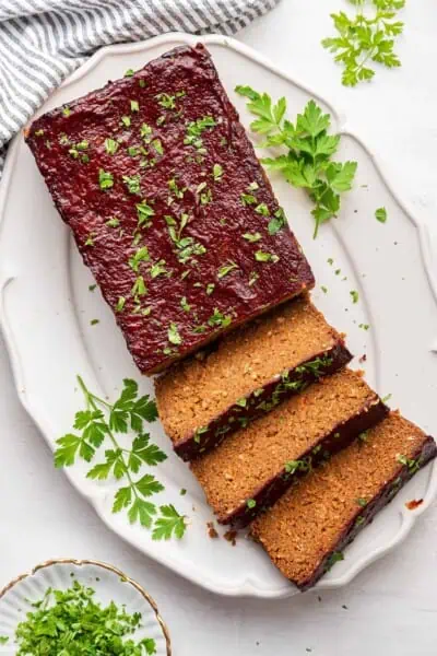 A serving tray with half a vegan meatloaf on it, uncut, and three slices of vegan meatloaf, topped with parsley, with parsley on the serving tray